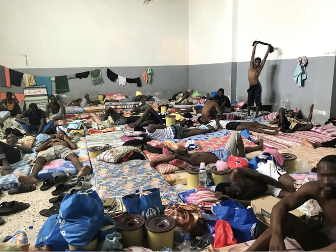 A large, crowded indoor room with many adult men lying on thin mattresses spread across the floor. Personal belongings, plastic containers, and bags are scattered around. Clothes hang along the walls. Most individuals are resting or lying down, while one man stands stretching with his arms raised. The setting appears crowded, with limited space between sleeping areas and minimal furnishings.