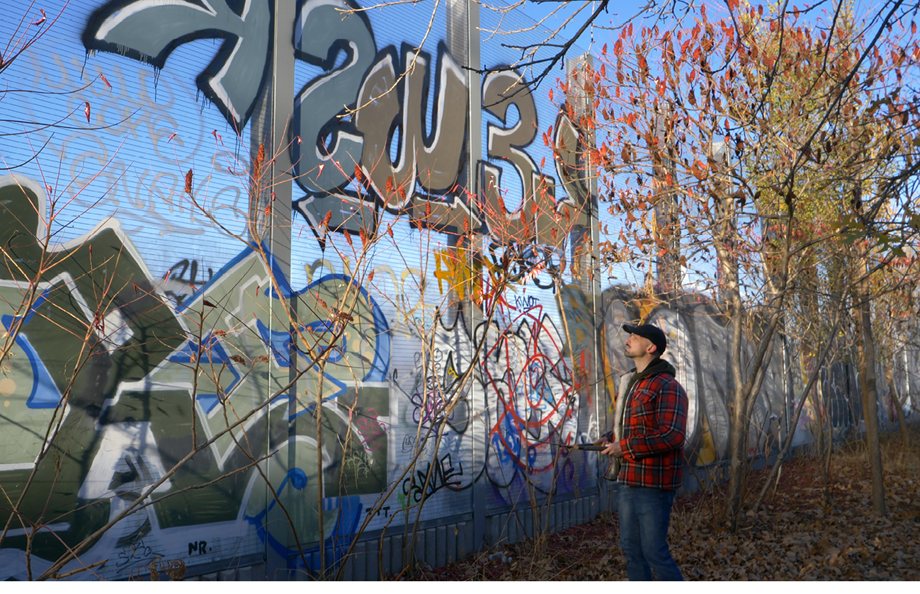 A person looks up at sumac trees and graffiti on an outdoor path.