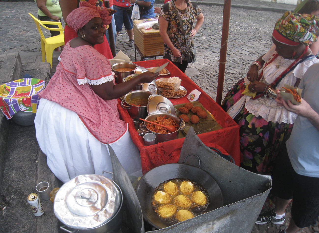 An Afro-Brazilian woman in traditional dress smiles as she serves acarajé, a savoury black-eyed pea fritter descended from West Africa, and other delicacies cooked in palm oil in the heart of the historic city centre of Salvador da Bahia, Brazil.
