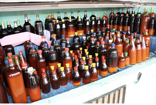 Various repurposed bottles of artisanal azeite de dendê, or palm oil produced traditionally with large dugout mortars and wooden pestles, sit for sale in a bustling outdoor market in Valença, Bahia.