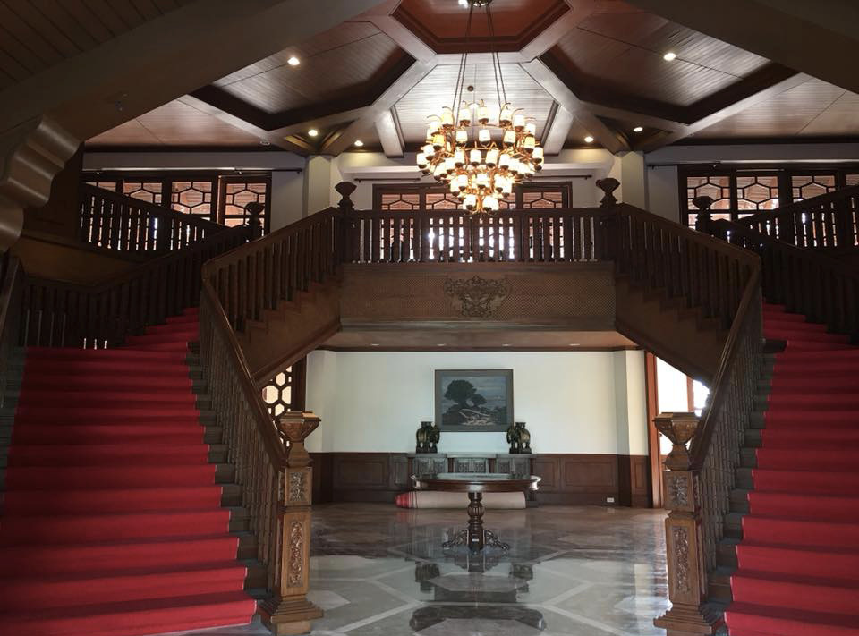 A photograph of the interior lobby of the Tahanang Pilipino. To the left and right are two wide, wooden staircases. Each is covered in red carpet. The railings and the ceiling are made of wood. A light-colored chandelier made of polished coconut shells hangs from the ceiling.