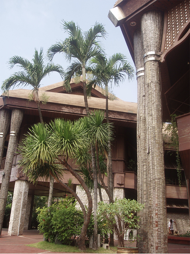 An exterior view of the Tahanang Pilipino. Stone pillars raise the wooden building above the ground. Four large and thick coconut tree trunks act as supports for the wooden roof. A thatch of six smaller coconut trees grows between the columns. In the background, a groundskeeper tends to a decorative raised garden bed.