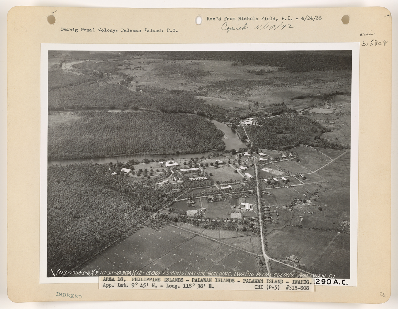 An aerial photograph of the Iwahig Penal Colony in black and white. The view is expansive. There are a handful of rectangular buildings in the centre. To the left is a large agroforest of coconut trees planted in neat rows and spaced at even distances. From the above, the forest appears to look like a grid. The Iwahig River is in the distance in the middle and top of the photograph. To the right and bottom are cleared fields separated by a road.