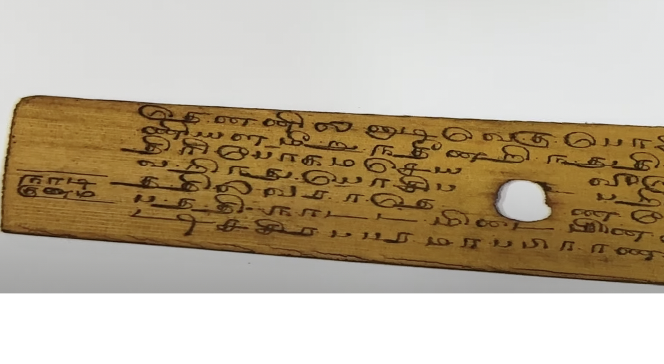 This image is a close-up view of one of the palm-leaf manuscripts that reveals finely etched Tamil characters darkened against the surface of the leaf. A circular binding hole interrupts the text near the centre, while the delicate grain of the palm leaf and the hand-drawn script convey the age and craftsmanship of the historical document