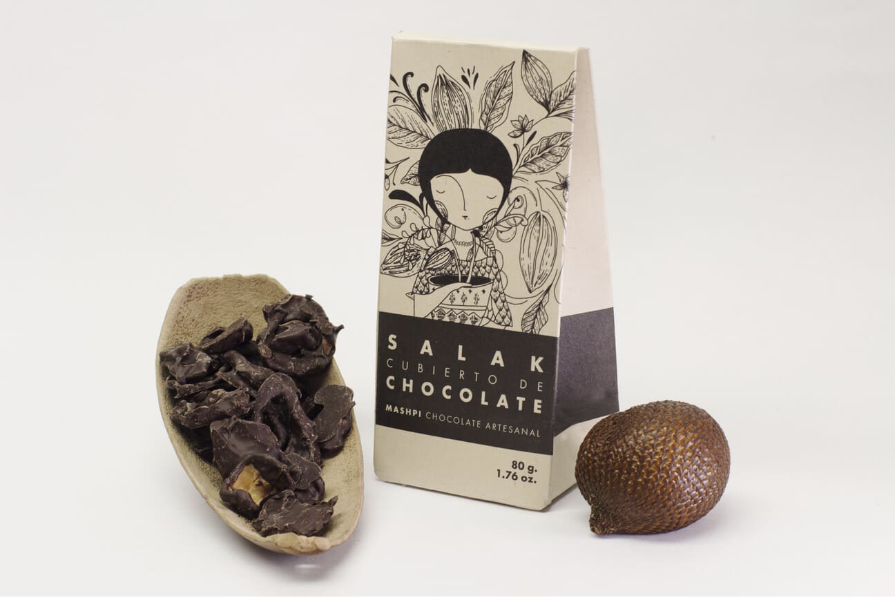 An image showing artisanal chocolate-covered Salak (snake fruit) by Mashpi Chocolate. On the left, a light-coloured, shallow bowl holds several dark, irregular pieces of chocolate-covered fruit. In the center is a vertical paper package featuring an illustration of a woman surrounded by cacao leaves and pods, with text reading ‘SALAK CUBIERTO DE CHOCOLATE’ and ‘MASHPI CHOCOLATE ARTESANAL’.To the right of the package sits a whole, unpeeled Salak fruit, identifiable by its brown, scale-like outer skin.
