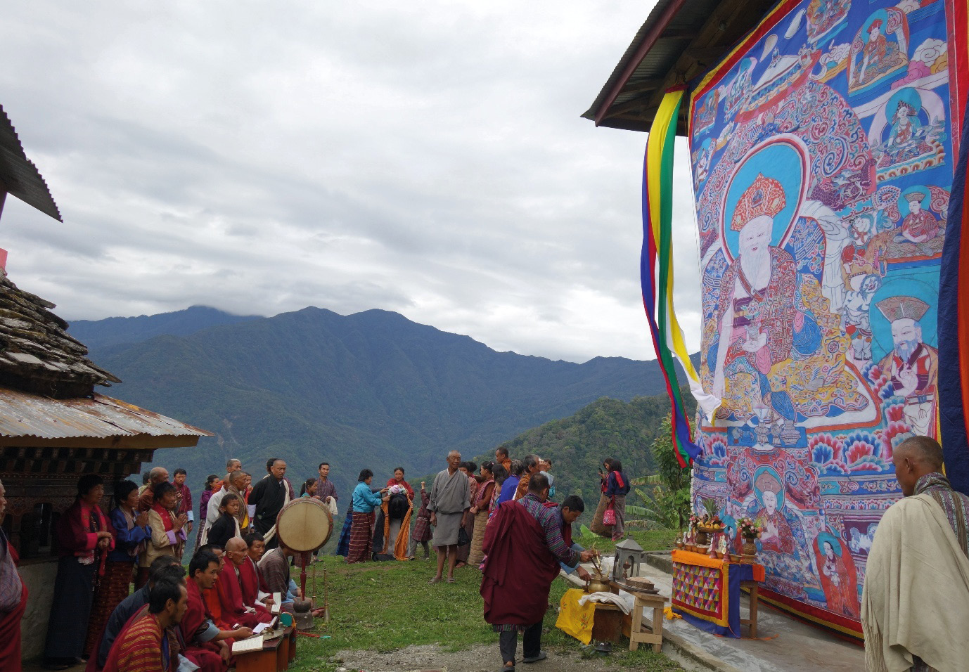 Priests performing a ritual in the temple lawn against the backdrop of surrounding mountains with a large mthong ’grol, appliqued Buddhist tapestry.