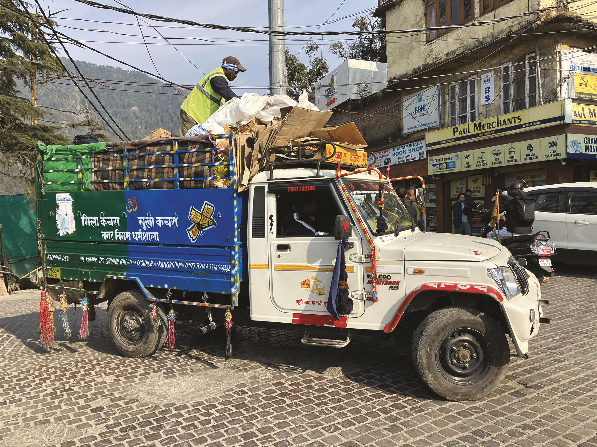 Photograph showing a stationary jeep, with a man wearing a high visibility vest standing atop rubbish loaded on to the open cargo area at the back.