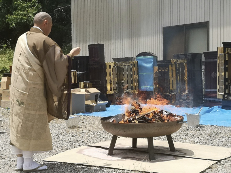 A Buddhist priest clad in light-brown ceremonial robes sprinkling consecrated water on small container with burning religious paraphernalia. Behind, Buddha altars are lined up in front of building.