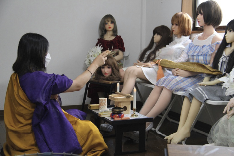 The priest sits before six human-like dolls, raising her right hand in a ritual gesture directed toward them and behind a small table, upon which rest a sutra book, a bell and two lit candles.