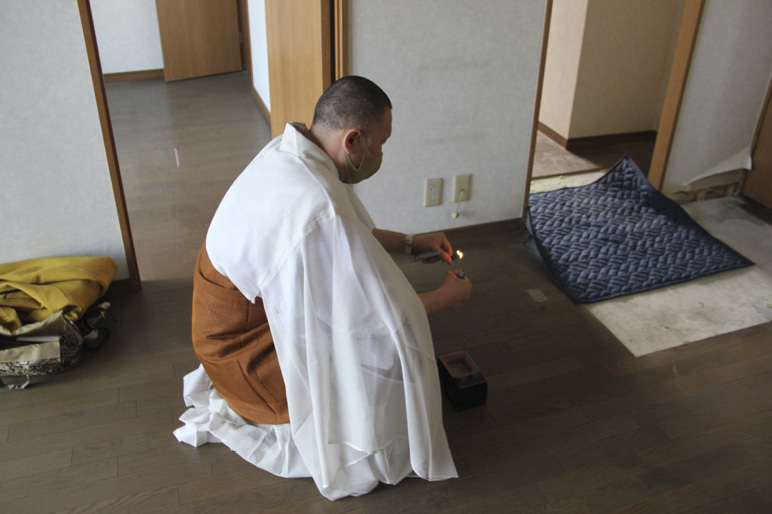 A priest clad in white and brown robes sitting on the floor in front of a small incense box, lighting an incense stick. Apart from a blanket and some of the priest’s belongings, the rooms are all empty.
