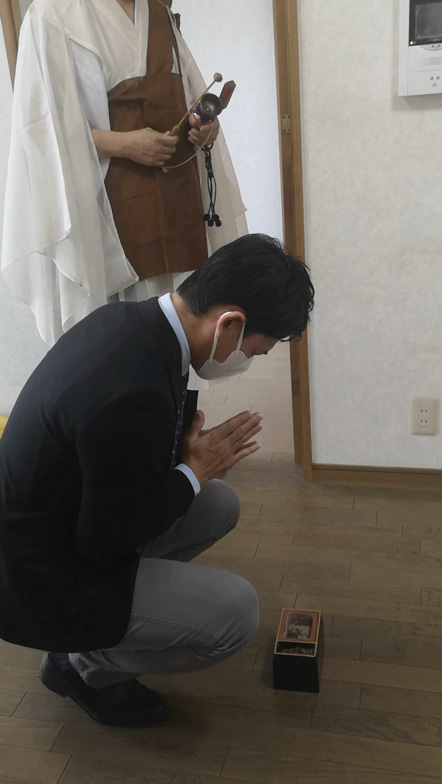A young man, formally dressed in a suit and wearing a face mask, kneels before a small incense box. Behind him, the priest stands holding a bell and the wooden striker used to ring it.